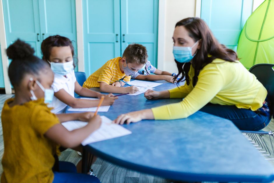 Teacher and children wearing masks engage in a classroom activity, emphasizing safety and learning.