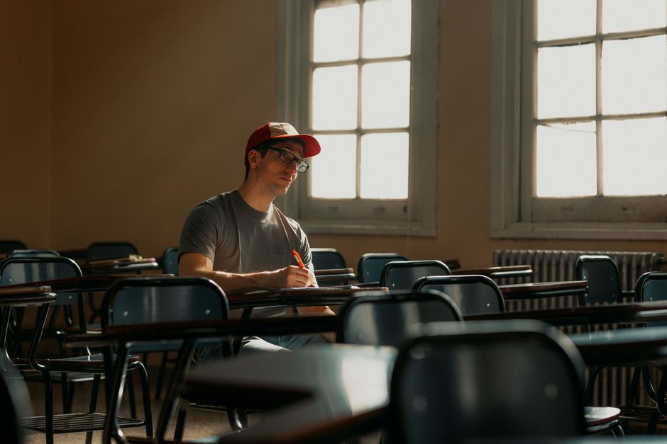 A university student in a cap studying alone in an empty classroom, lit by natural sunlight.